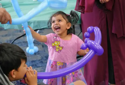 Smiling young girl in a pink dress plays with blue balloon animals at a party, a boy nearby watching the fun.