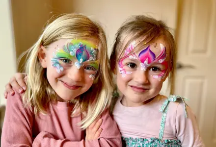 Two young girls indoors with butterfly face paint, smiling and standing close with arms around each other.