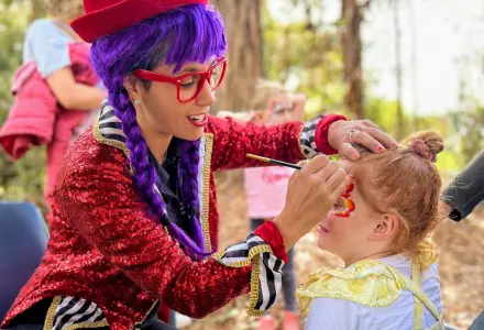 Clown with purple hair and red glasses applies face paint to a young girl outdoors.
