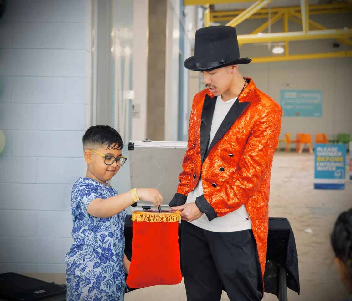 Magician in a sparkly red jacket and black top hat performs a trick for a smiling young boy with glasses as they handle a red velvet prop bag, in a bright indoor setting.
