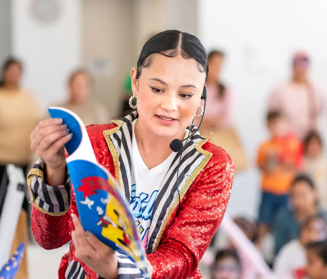 Performer in a red sequined jacket with a headset presents a curved blue prop to a crowd at a children’s event.