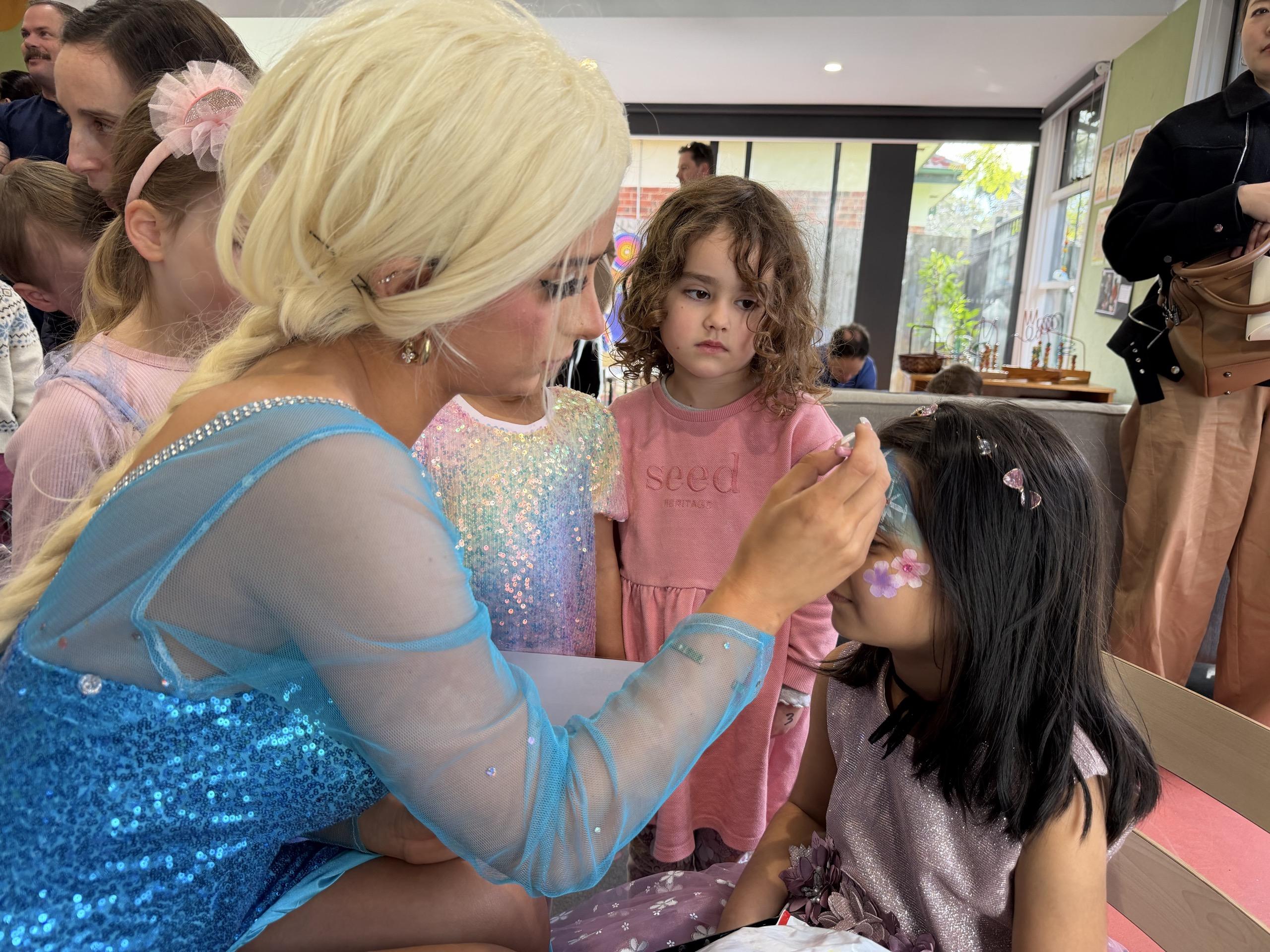 IMG_0451 A woman in a blue sequined costume interacts with several young girls at a party or event, helping with a craft or activity.