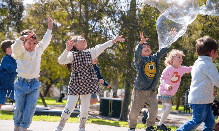 Recommended Party Supliers – 3 bg (1) Group of children playing outdoors, jumping with arms raised as a large bubble floats above them on a sunny day in the park.