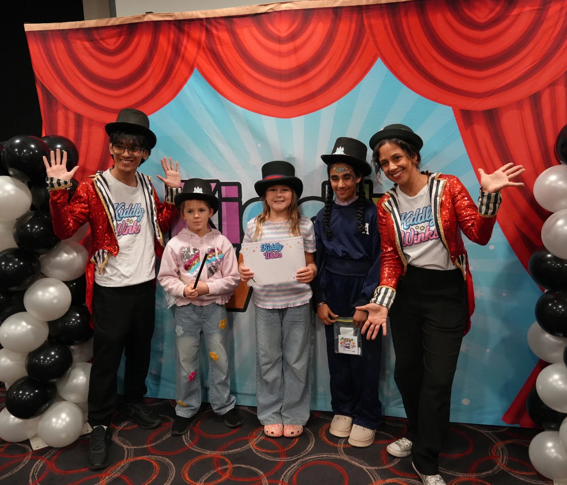 Group of five children and performers posing in front of a colorful backdrop with red curtains and balloons; one child holds a certificate.
