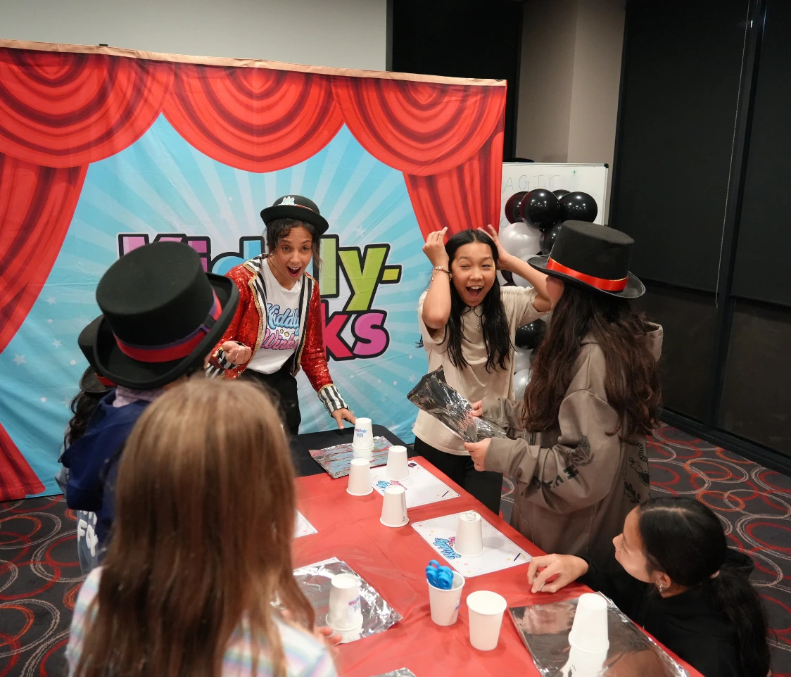 Children at a birthday party watch a performer in a red sequined jacket and top hat near a blue stage backdrop with red curtains.