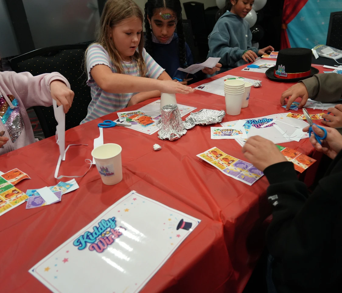 Children sit around a red table doing arts and crafts with scissors, paper, and foil at a party.