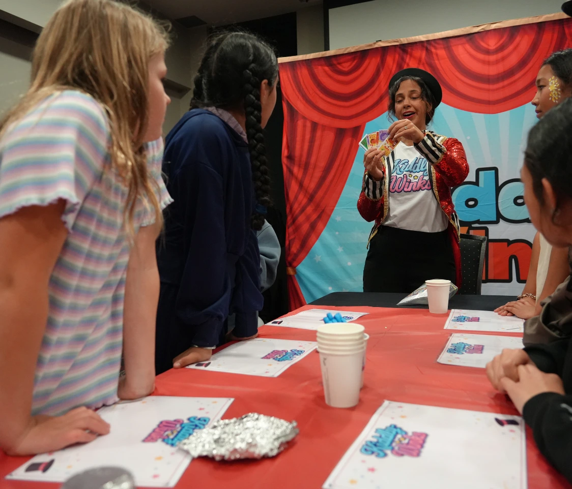 Performer in a red sequined jacket shows a fan of playing cards to kids at a table with a red tablecloth.