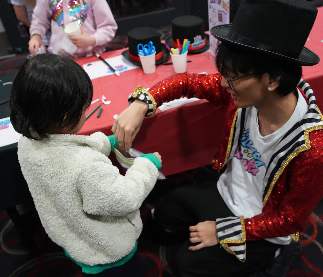 Magician in a red sequined jacket and top hat performs a trick for a child at a red craft table
