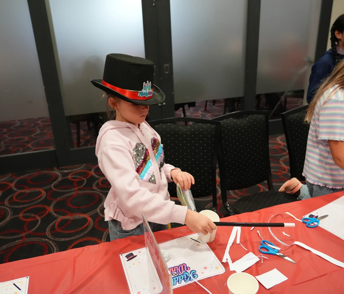 Girl in a pink hoodie wearing a black top hat with a red band, crafting with scissors and paper at a red table.