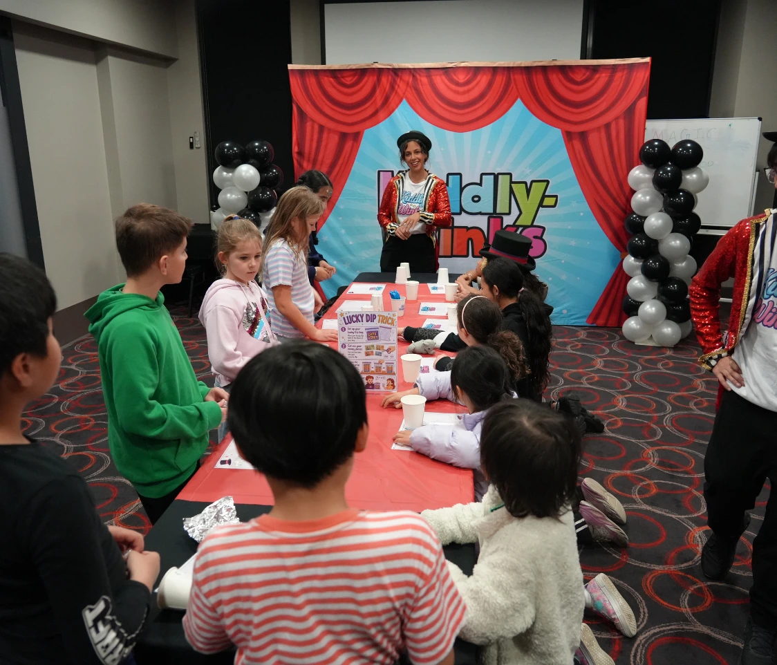Children line up around a long red-tablecloth booth at a kids' party or show, watching a performer in a red sequined jacket on a bright blue stage with red curtains and balloons nearby.