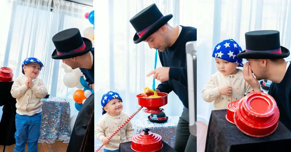 Magician in a black top hat performs a birthday magic trick for a toddler wearing a blue star bandana, with balloons nearby.