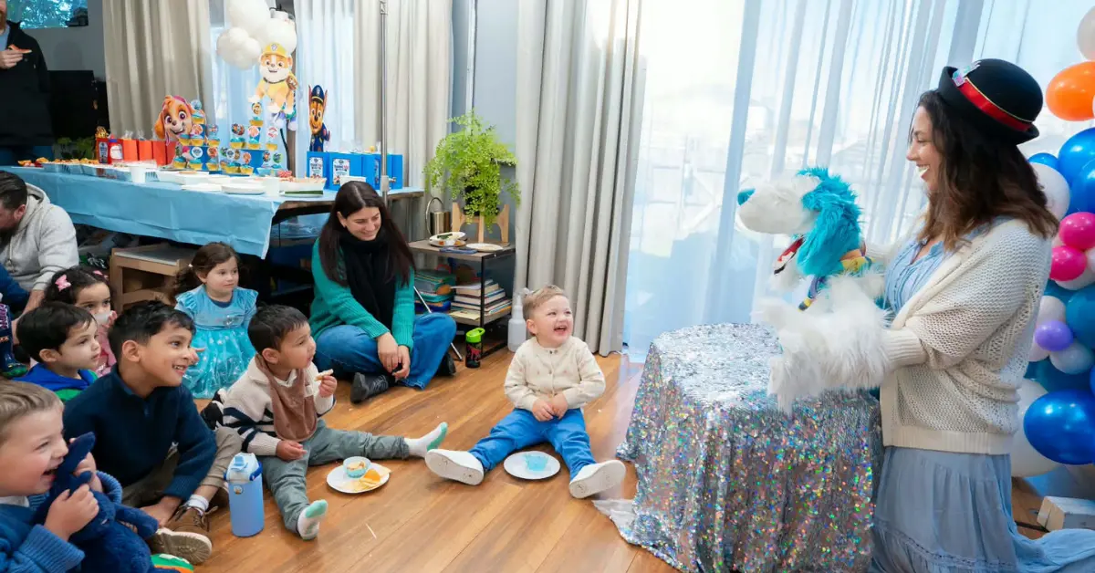 Children sit on the floor laughing as a performer uses a white puppet dragon during a birthday party, with a glittery table and balloons nearby.
